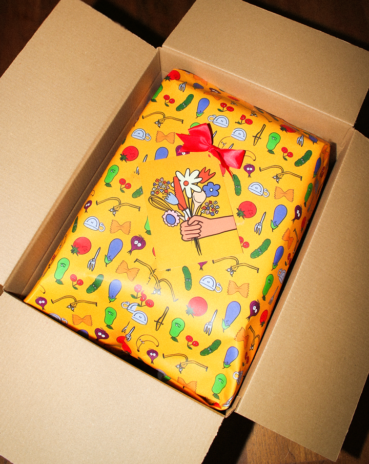 A high-angle view of a cardboard shipping box containing a gift-wrapped item. The premium yellow gift wrap features a playful illustrated pattern of vegetables (cucumber, carrot, tomato) and a red bow with a 'Thinking of you' gift card. A ready-to-gift Montessori set