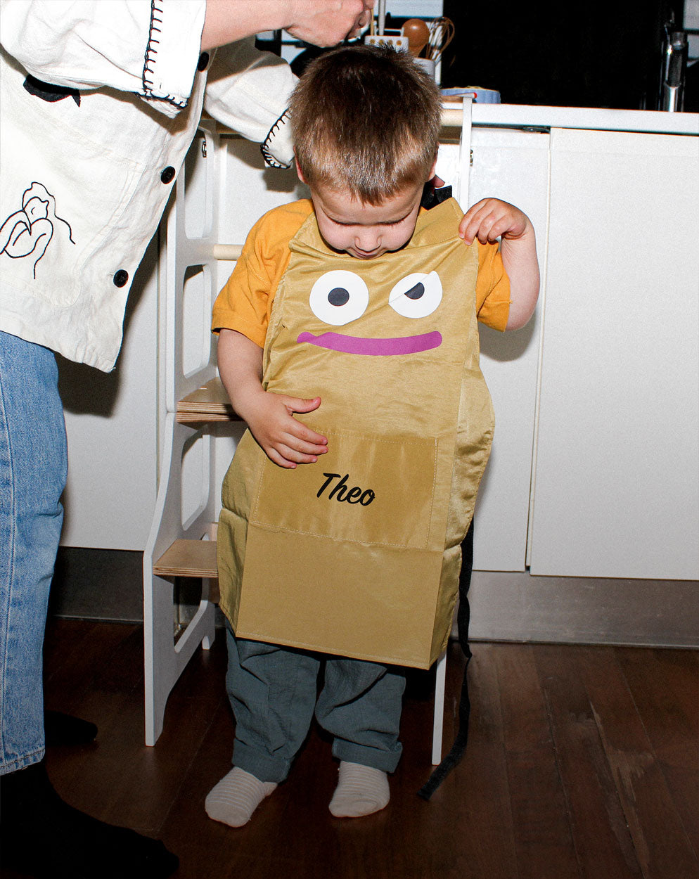 A toddler boy putting on his Kindy™ personalised apron featuring a friendly character face and the name 'Theo' printed on the front. Highlights the excitement of starting a Montessori practical life kitchen activity.