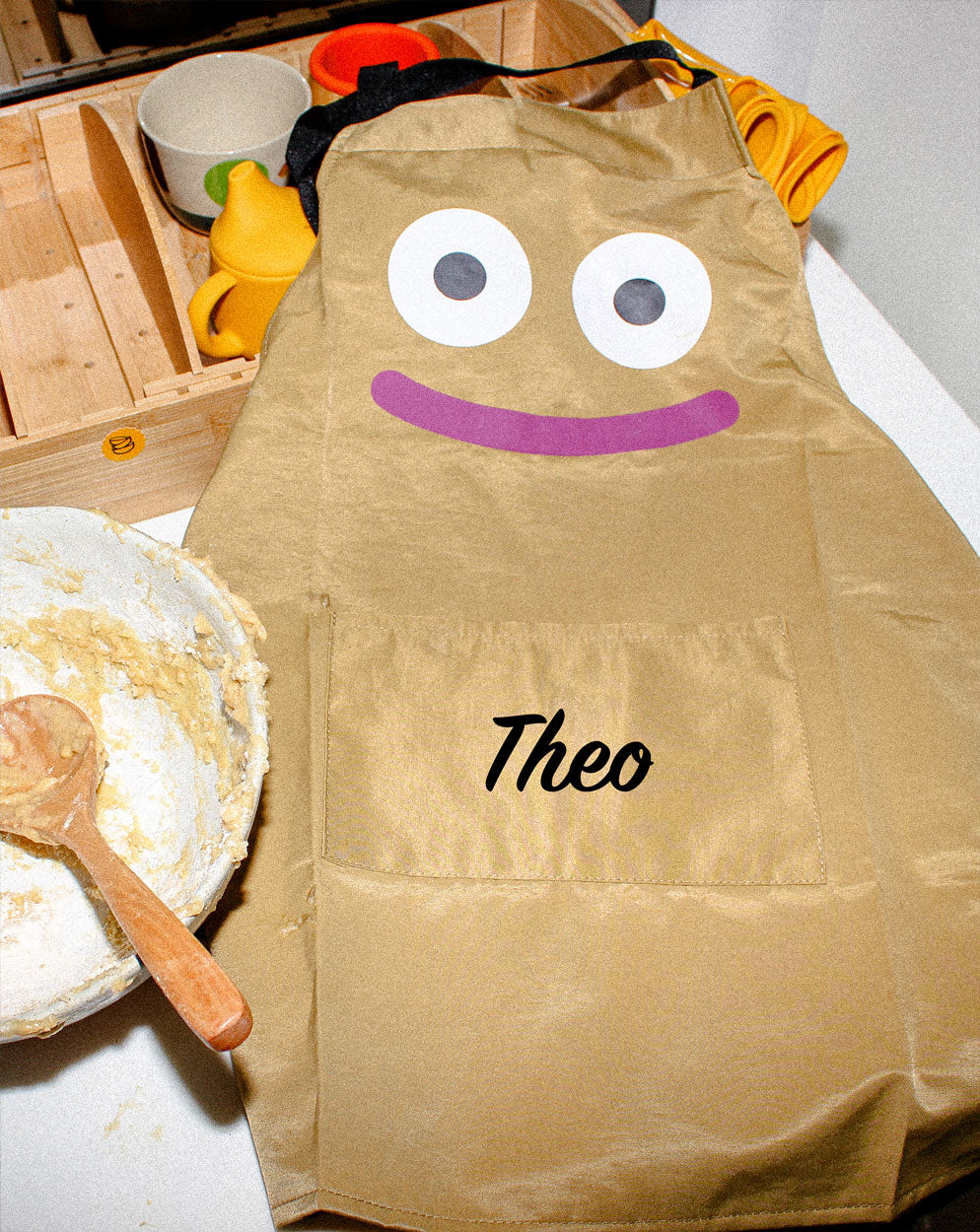 Close-up of the Kindy™ child's kitchen apron in a neutral sand colour, personalised with the name 'Theo.' The apron features an playful character design with eyes and a smile, encouraging kids to get creative and messy in the kitchen